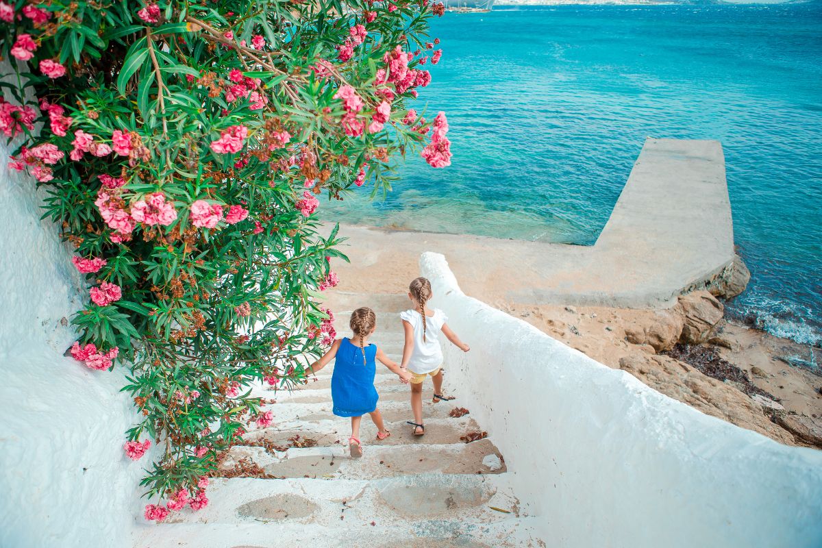 Two young girls holding hands, walking down a whitewashed staircase adorned with vibrant pink flowers, towards a serene, turquoise sea.