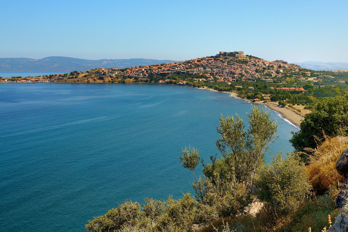 A scenic coastal town with a hilltop castle overlooking a bay, featuring blue waters and a shoreline with greenery in the foreground.