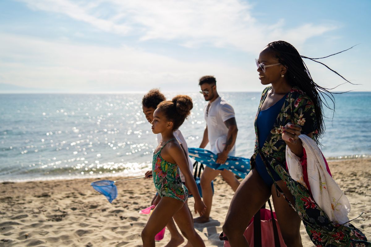 A family walking along a sunny beach, with two children in swimsuits and adults carrying beach gear and bags. The sea is calm and shimmering in the background.