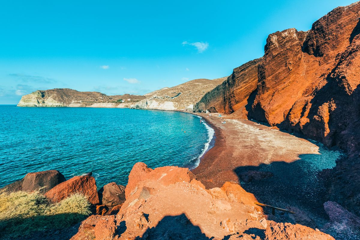 A picturesque beach with striking red cliffs, clear blue waters, and a small sandy shoreline. The landscape is rugged and scenic under a bright blue sky.