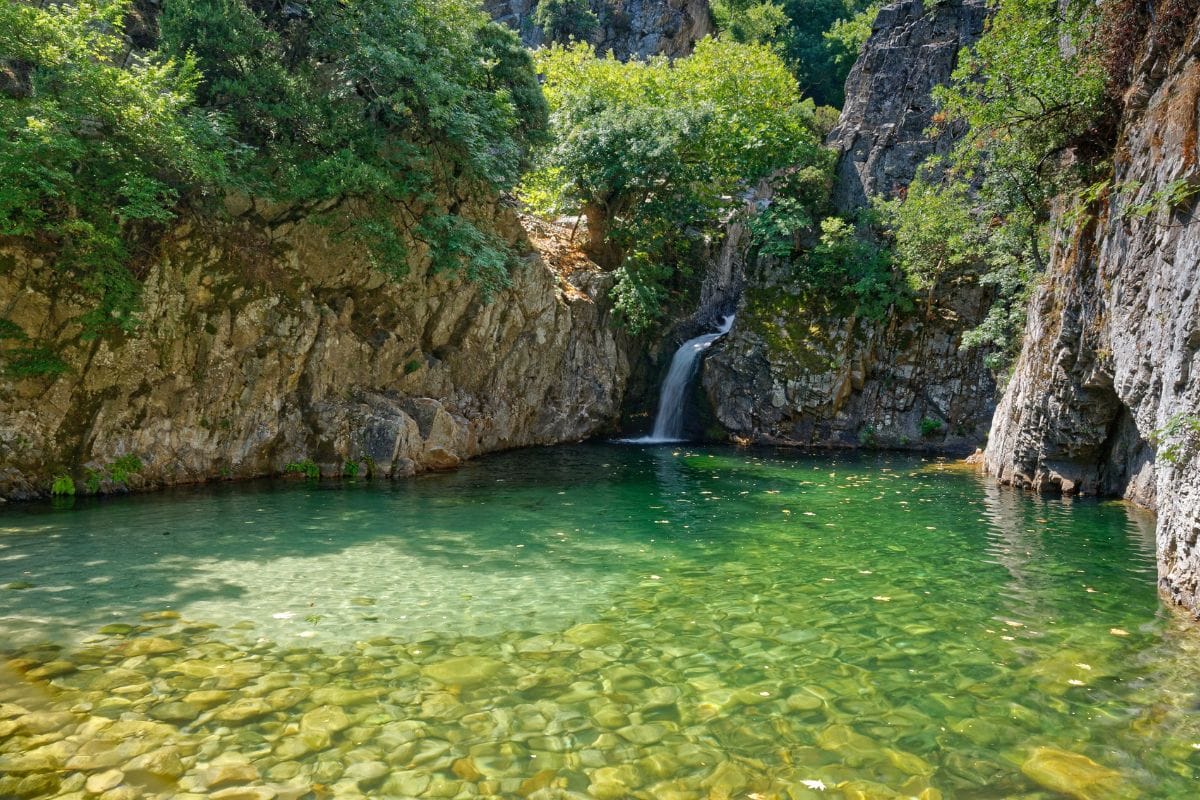 A serene natural pool with clear green water surrounded by rocky cliffs and lush vegetation. A small waterfall flows into the pool, adding to the tranquil atmosphere.