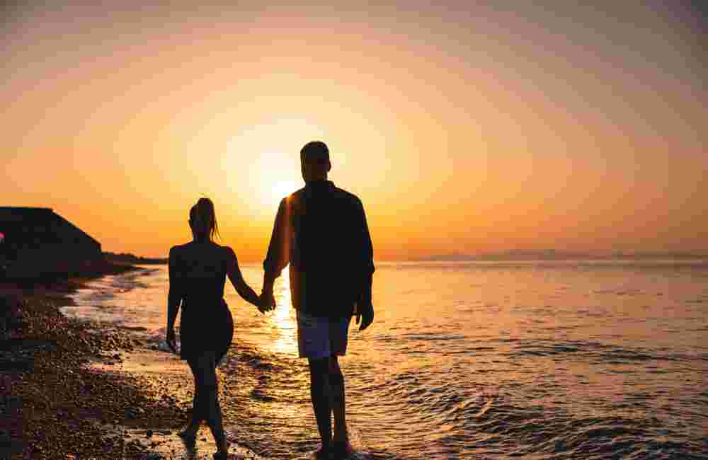 Silhouette Of A Romantic Couple Holding Hands While Walking On The Beach Towards The Sunset