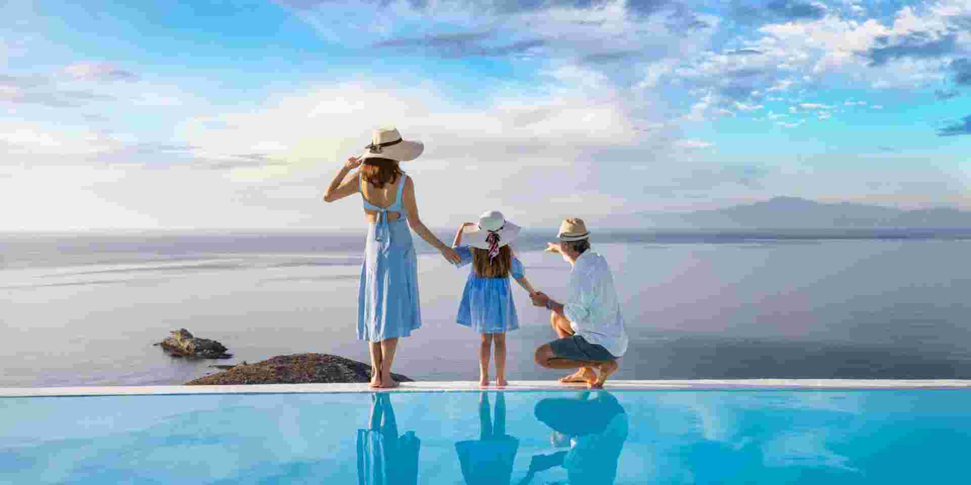 A beautiful family stands at the edge of a swimming pool and enjoys the view over the ocean during their holidays Greece family vacation_feature