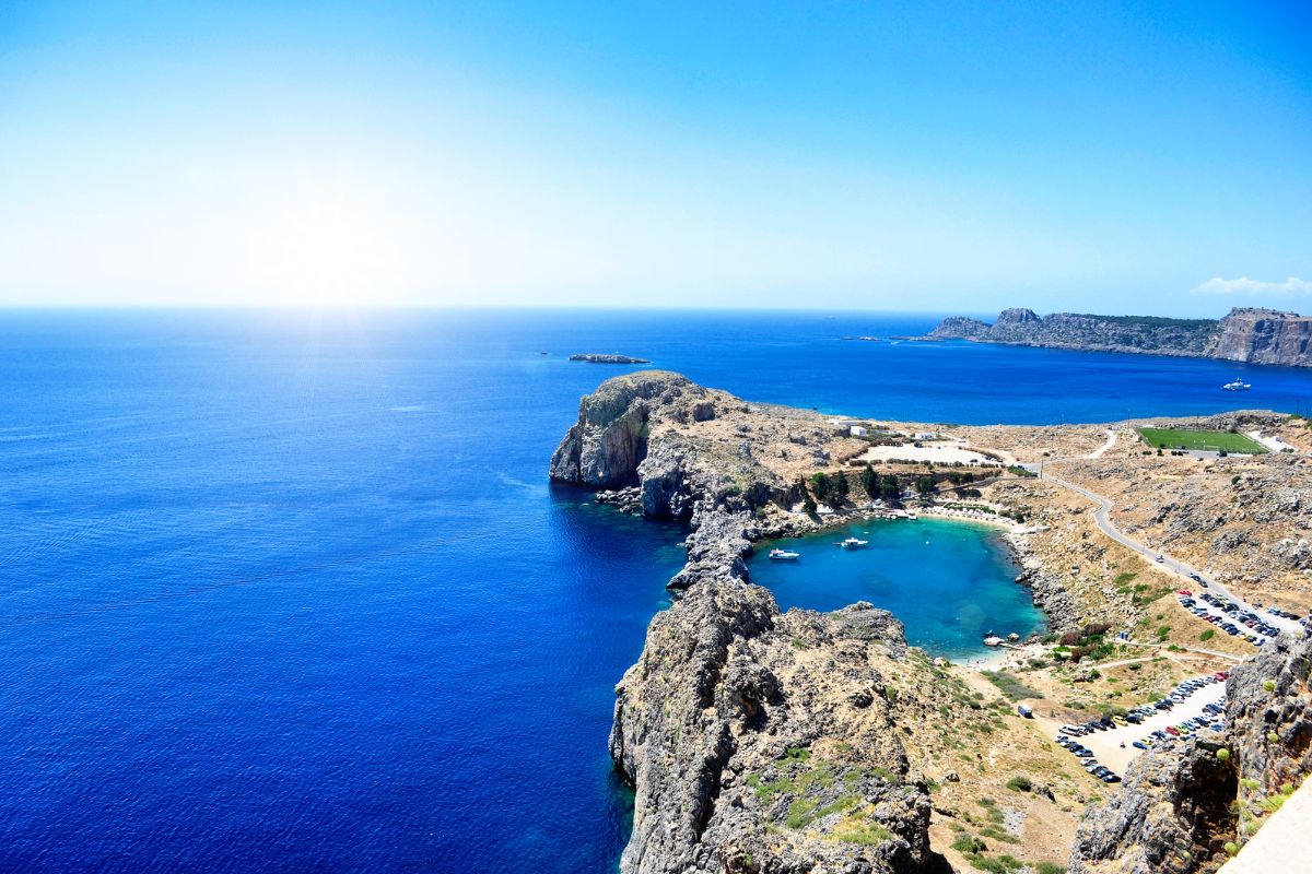 A breathtaking view of the Acropolis of Lindos in Rhodes, showcasing the ancient ruins perched on a rocky hill overlooking the deep blue Aegean Sea, with a small bay and scattered boats below.