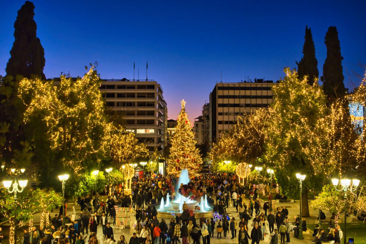 Syntagma Square in Athens is beautifully decorated for Christmas, with a large, illuminated Christmas tree at its center and twinkling lights adorning the surrounding trees. The square is bustling with people enjoying the festive atmosphere, with some gathered around a brightly lit fountain. The scene captures the vibrant holiday spirit against the backdrop of modern buildings under a twilight sky. 