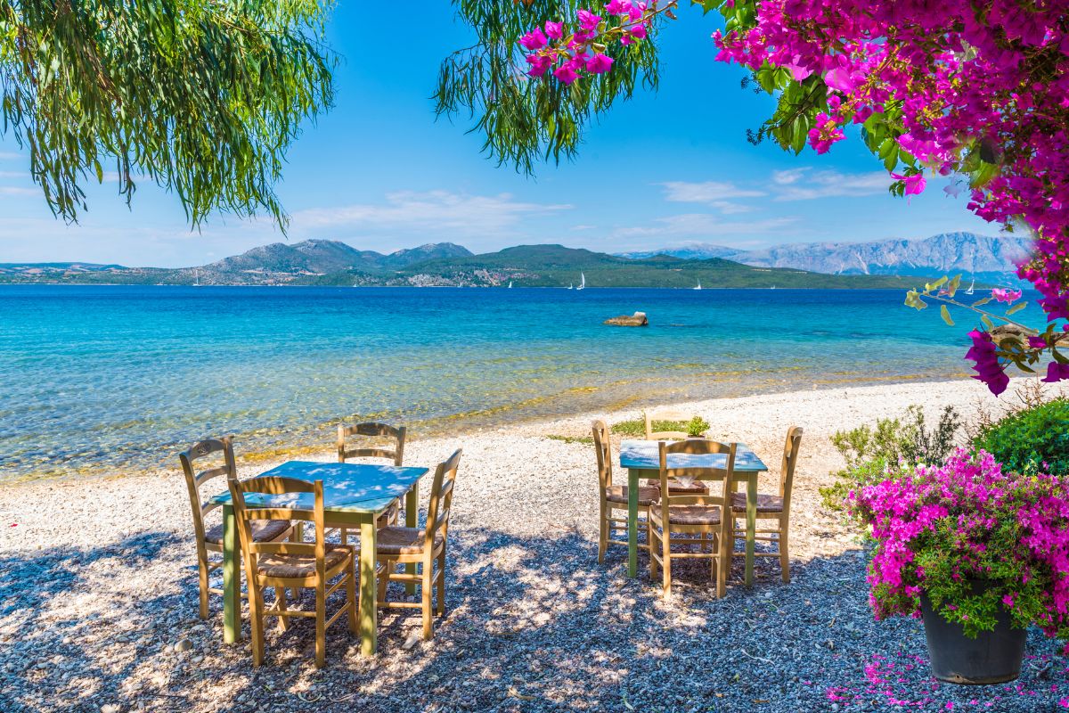 Beachfront taverna on Lefkada island, with wooden tables and chairs on a pebbled shore, overlooking the blue sea and distant mountains, framed by pink flowers and green foliage.