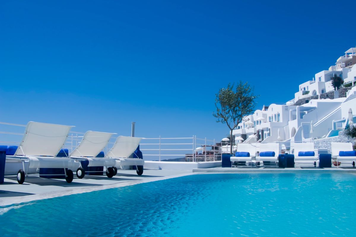 A luxurious hotel in Greece with white buildings, blue accents, and a serene pool area featuring lounge chairs under a clear blue sky.