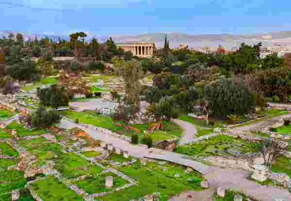 This stunning aerial photograph showcases the Temple of Hephaestus, one of the best-preserved ancient Greek temples, located in the Ancient Agora of Athens, Greece. Built in the 5th century BC, the temple is dedicated to Hephaestus, the god of fire and craftsmanship. The image captures the temple's majestic Doric columns standing amidst lush greenery and the remains of ancient structures. In the background, the cityscape of Athens and distant mountains create a striking contrast between ancient and modern times. This historical landmark is a key site for travelers, archaeologists, and history enthusiasts. ancient agora athens_what to see in greece_feature