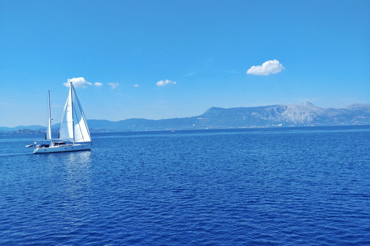 A sailboat glides across the calm, blue waters of the Ionian Sea with distant mountains under a clear, blue sky.