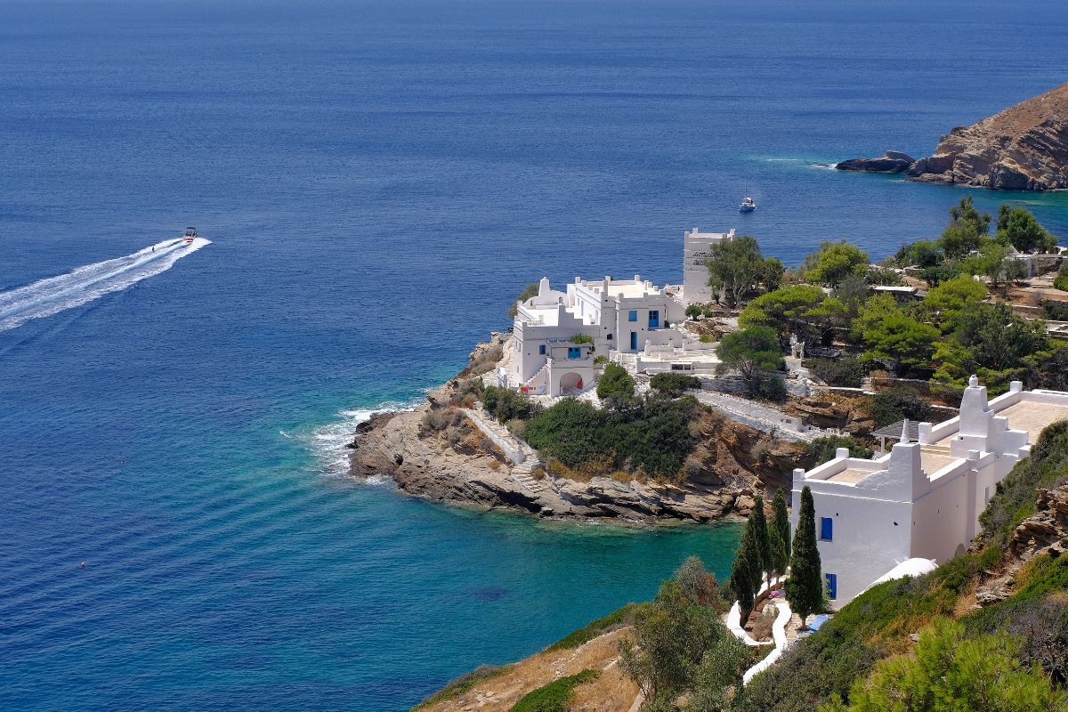 White buildings with blue accents are perched on the rocky coastline of Ios Island, Greece, overlooking the clear blue sea with a boat leaving a white trail in the water.