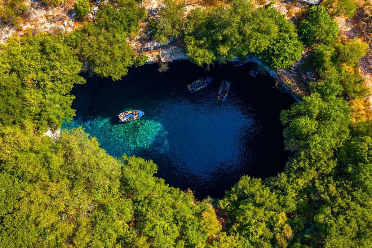 Aerial view of boats floating on the turquoise waters inside Melissani Cave, surrounded by dense green foliage in Kefalonia.