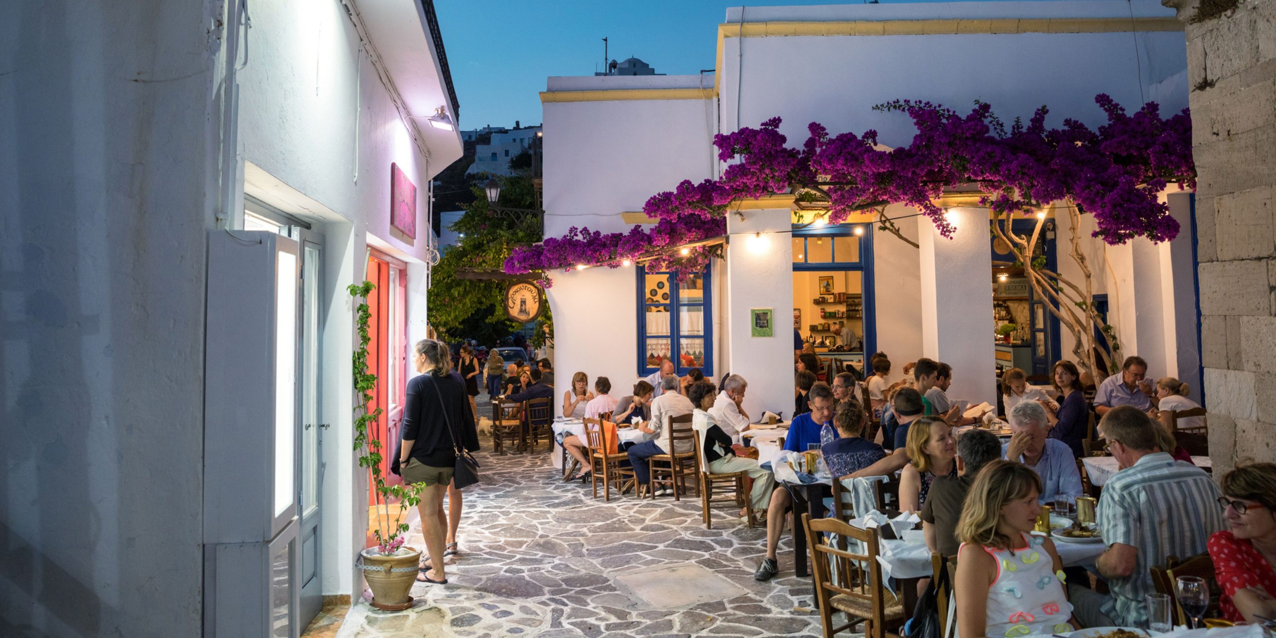 Milos island, Greece - June 11, 2018: Sunset scene in Plaka village which is full of shops and restaurants. People are dining in one of the restaurants decorated with bougainvillea flowers.