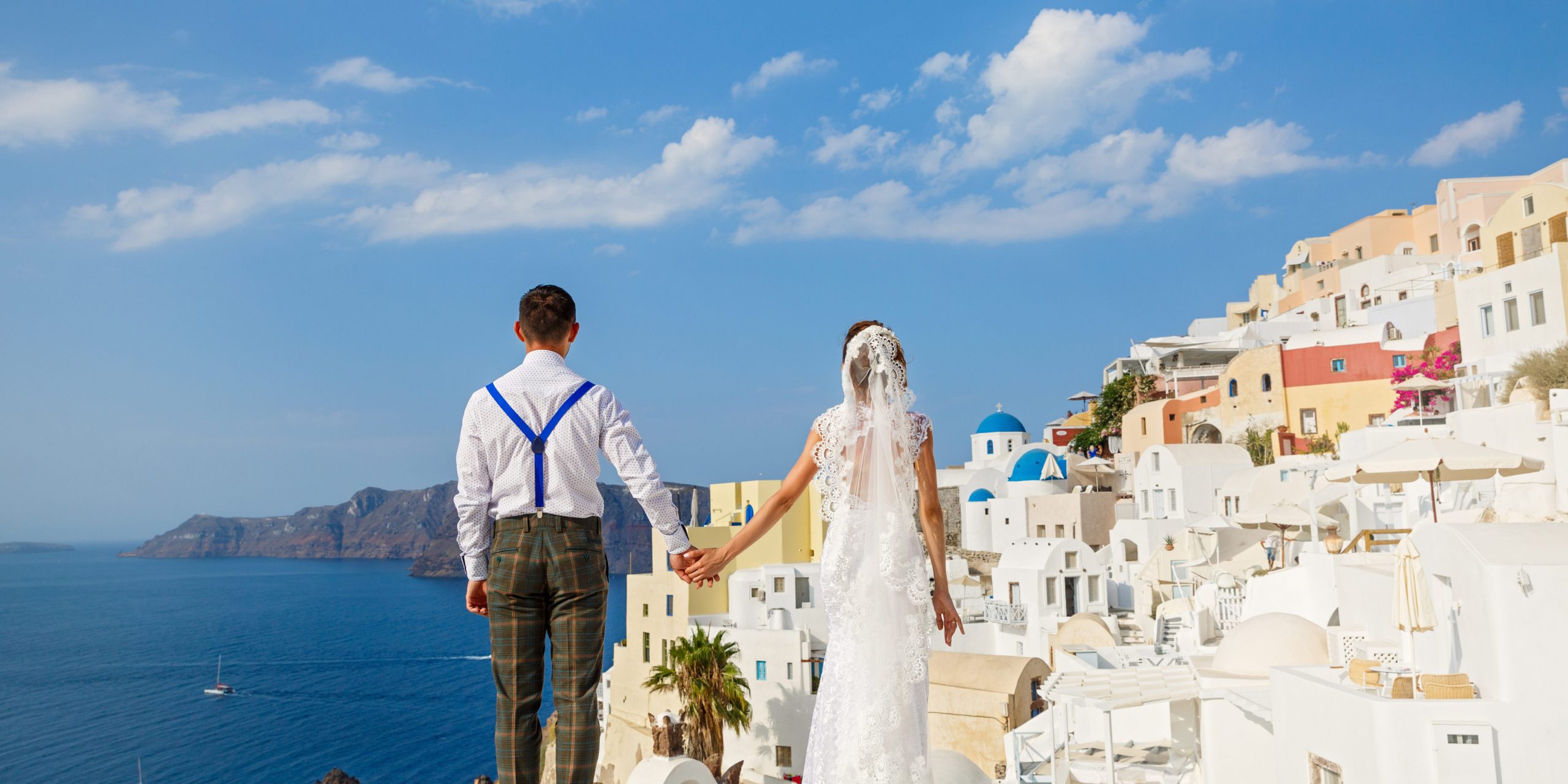 A couple in wedding attire holding hands, standing in front of the picturesque white-washed buildings and blue domes of Santorini, with the sea and mountains in the background.