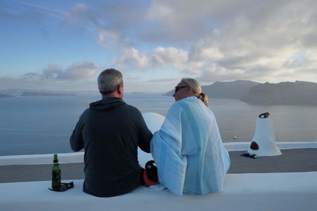 A senior couple sits together in Santorini, looking out over the sea and distant cliffs.