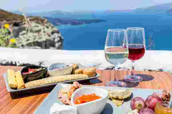 A picturesque setting in Santorini with two glasses of wine and a platter of assorted snacks, overlooking the stunning blue sea. wine tasting santorini_honeymoon