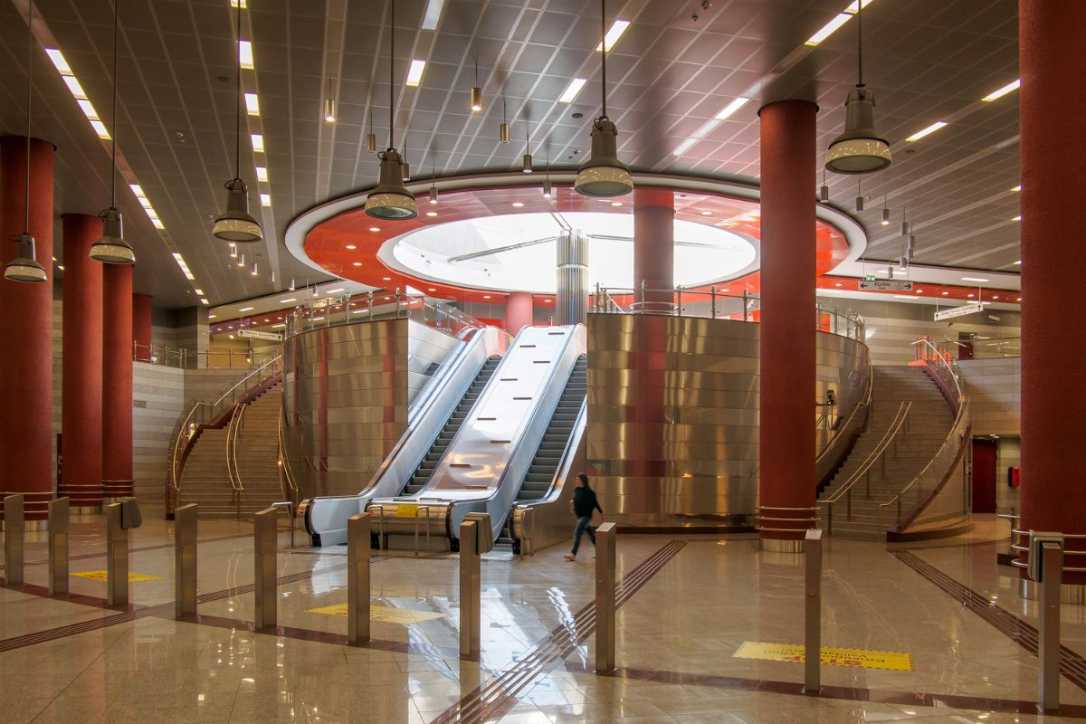 The image shows the interior of an Athens metro station, featuring modern escalators, curved staircases, and bold red pillars. The station has a sleek, contemporary design with well-lit spaces and a polished floor, reflecting the efficient and organized infrastructure of the city's public transport system.