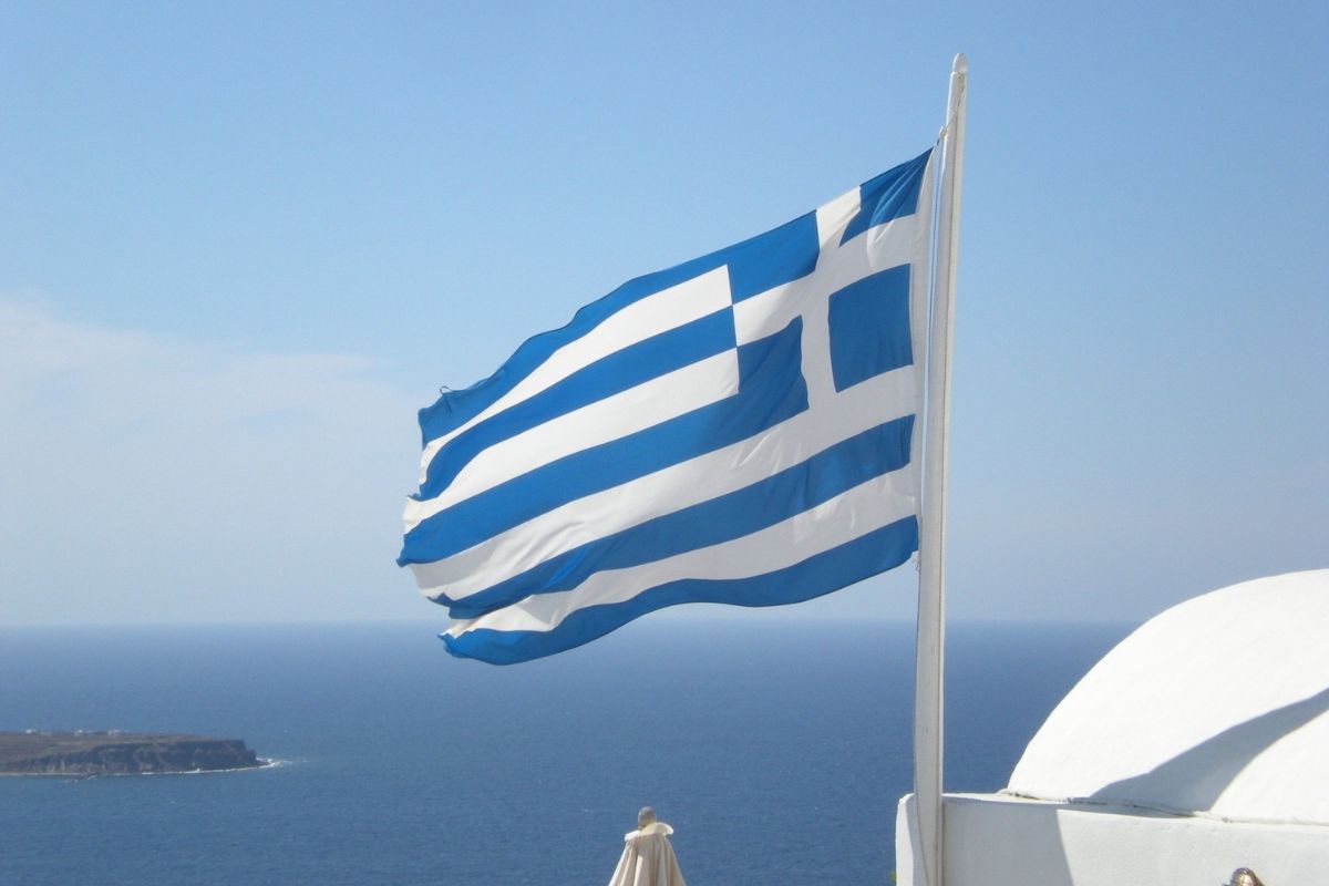 The image shows a Greek flag waving in the wind with a backdrop of a calm sea and distant coastline under a clear blue sky. The scene evokes a sense of tranquility and national pride.