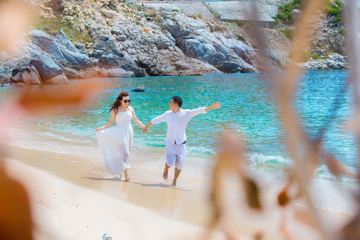 A couple in white attire walks hand-in-hand by clear turquoise water, with rocky cliffs in the background.