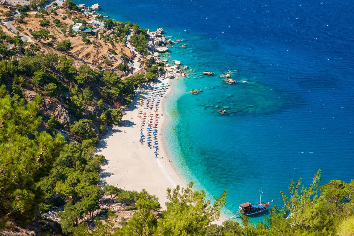 The image shows a picturesque beach on Karpathos Island, featuring a stretch of white sand bordered by turquoise waters. Lush greenery and rocky outcrops surround the beach, with colorful umbrellas and a boat anchored near the shore, highlighting the idyllic and serene coastal scenery.