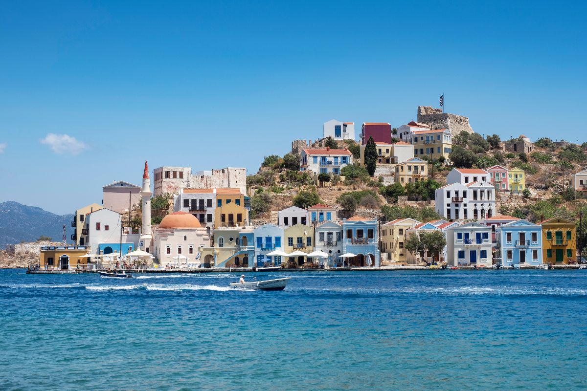 The image shows the picturesque waterfront of Kastellorizo Island, featuring a row of colorful, traditional houses nestled along the shore. The buildings are set against a backdrop of hilly terrain, with a clear blue sky above and calm waters in the foreground, reflecting the island's serene and charming atmosphere.