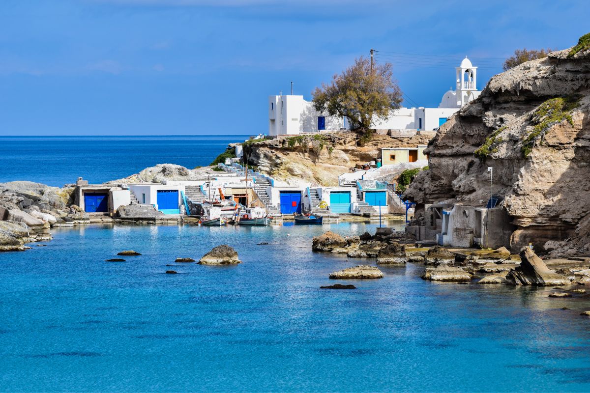 A serene coastal scene on Milos Island, featuring turquoise waters, traditional white buildings, and small fishing boats docked near rocky cliffs.