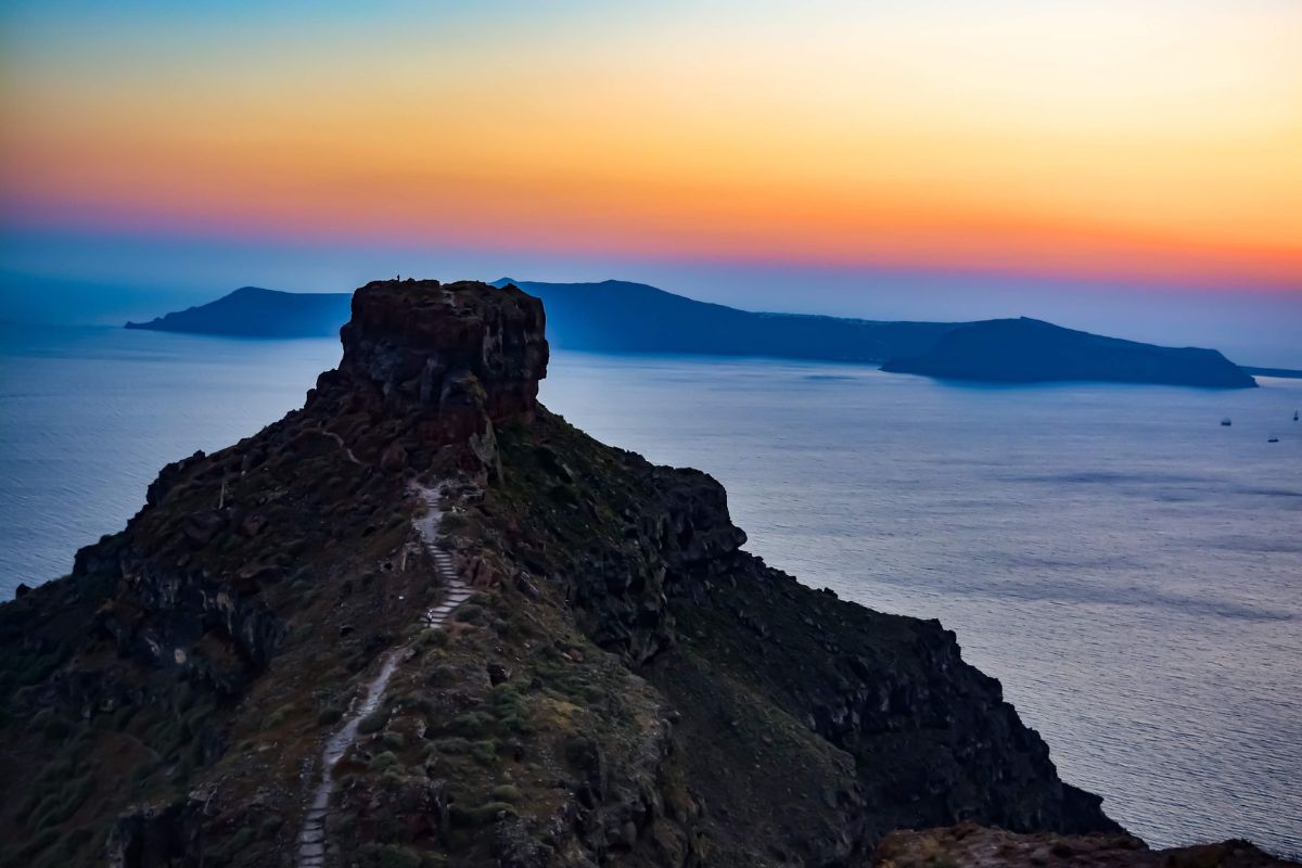 A view of Skaros Rock in Santorini at sunset, with the rugged rock formation silhouetted against a colorful sky and the calm Aegean Sea in the background.