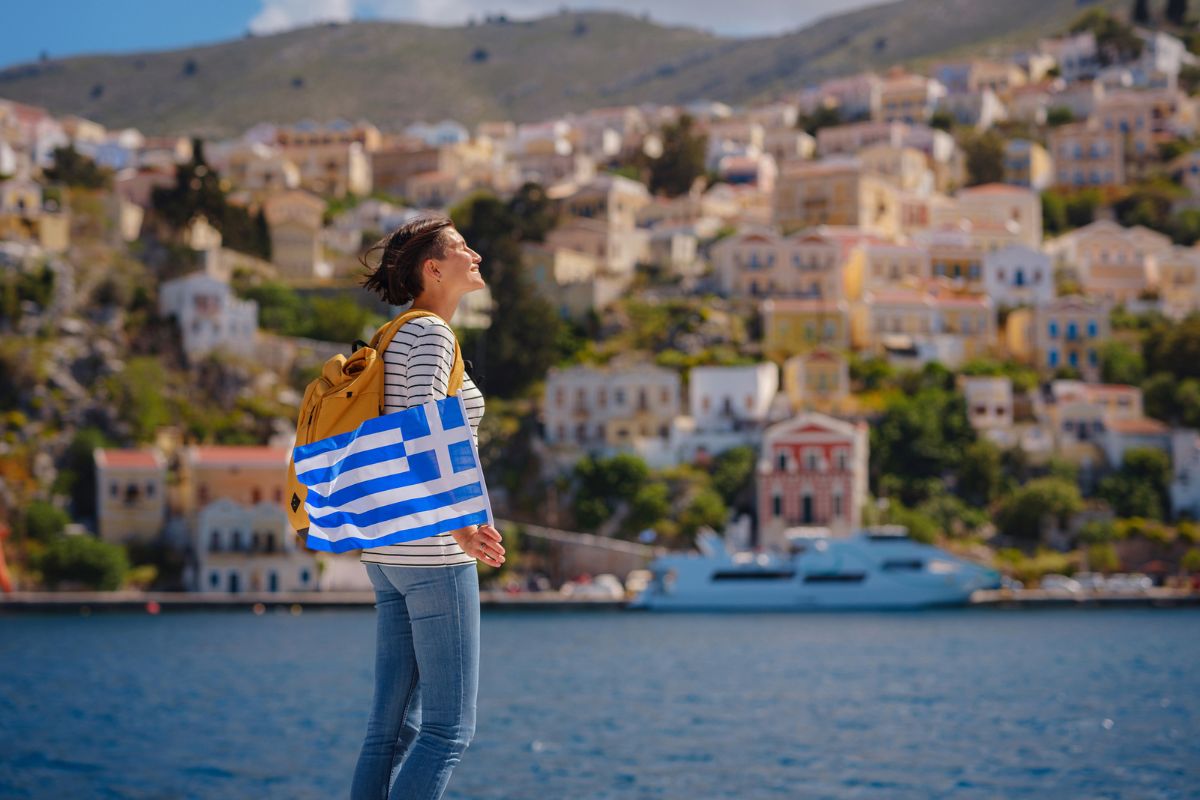 Nice asian Happy Female with backpack anf greek flag Enjoying her holidays on Symi Islands. View of port Symi or Simi, is tiny island of Dodecanese, Greece, calm atmosphere and fabulous architecture.