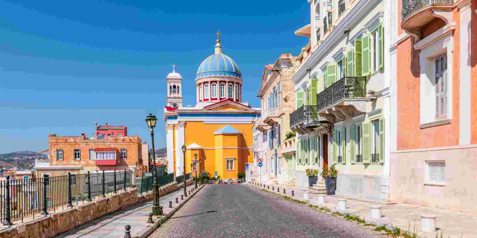 A street view in Ermoupolis, Syros, featuring colorful buildings and a church with a blue dome under a clear sky. ermoupolis syros_accessible greek islands for seniors_feature