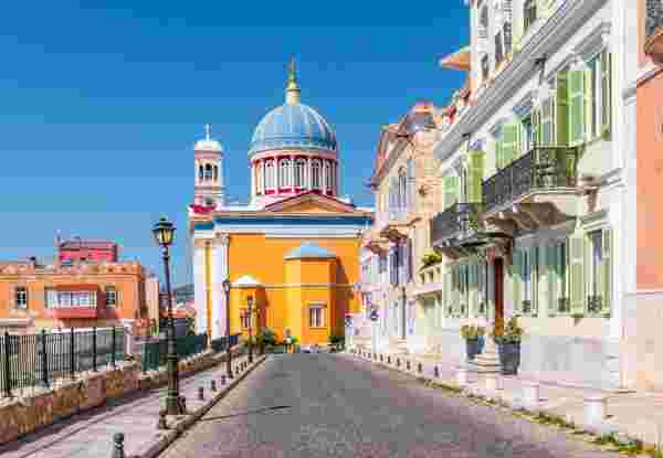 A street view in Ermoupolis, Syros, featuring colorful buildings and a church with a blue dome under a clear sky. ermoupolis syros_accessible greek islands for seniors_feature