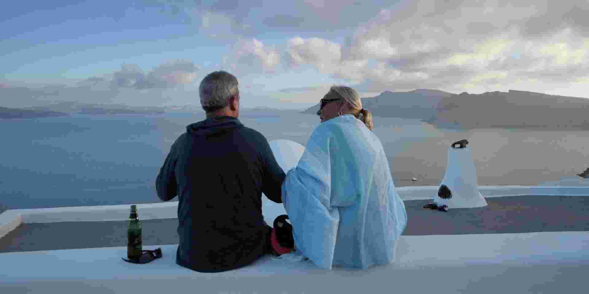 Santorini, Greece - 25 May, 2019: Senior couple travelers enjoy Santorini summer vacation sitting on wall having a beer drink and looking at amazing panoramic view over the roofs santorini_greek island for older couples_feature