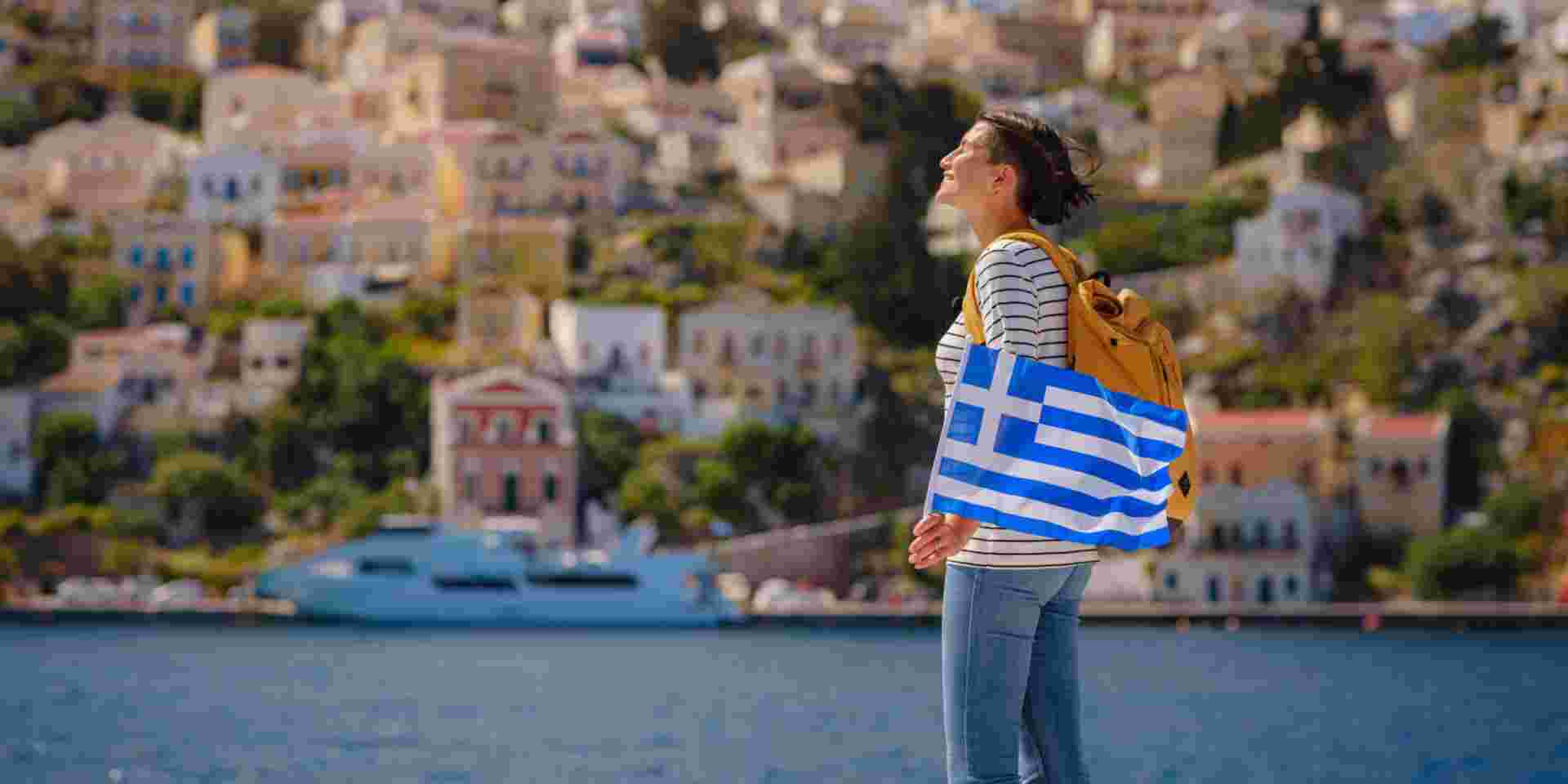 Nice asian Happy Female with backpack anf greek flag Enjoying her holidays on Symi Islands. View of port Symi or Simi, is tiny island of Dodecanese, Greece, calm atmosphere and fabulous architecture. symi island solo travel_is greece safe to travel to_feature