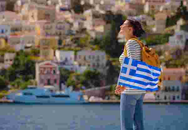 Nice asian Happy Female with backpack anf greek flag Enjoying her holidays on Symi Islands. View of port Symi or Simi, is tiny island of Dodecanese, Greece, calm atmosphere and fabulous architecture. symi island solo travel_is greece safe to travel to_feature