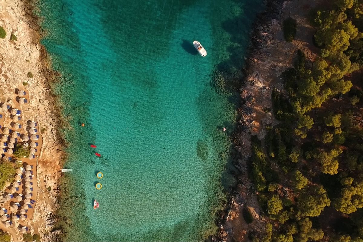 An aerial view of Agistri Island's clear turquoise waters, with a boat anchored near the rocky shoreline, surrounded by lush green trees and a small beach with sun umbrellas.