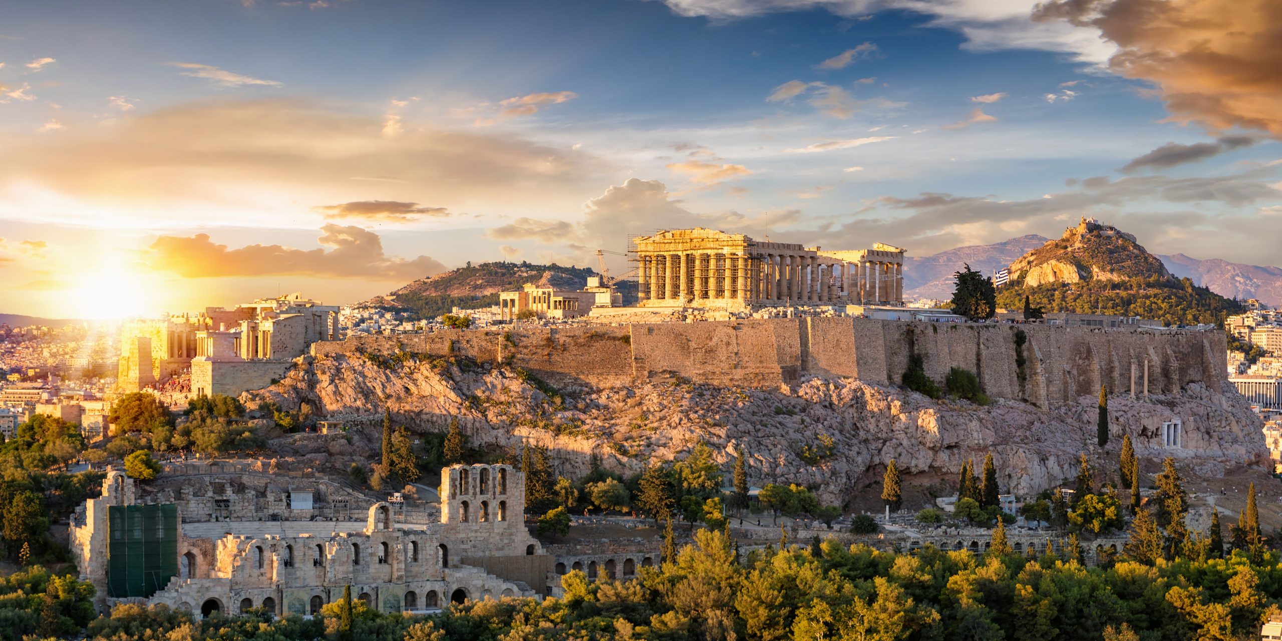 A stunning view of Athens at sunset, showcasing the Acropolis and its surrounding landmarks, with a warm glow highlighting the ancient ruins and the city below.
