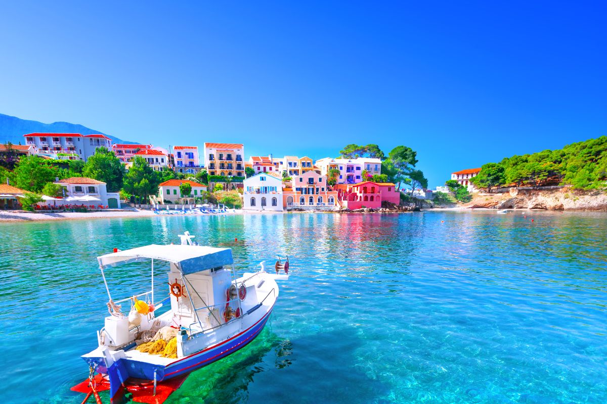 The image shows a colorful seaside village in Kefalonia, Greece. Vibrant houses in various pastel shades are nestled along the coastline, with crystal-clear turquoise water in the foreground. A small fishing boat floats calmly on the water, adding to the peaceful Mediterranean atmosphere. Lush greenery and bright blue skies complete the picturesque scene.
