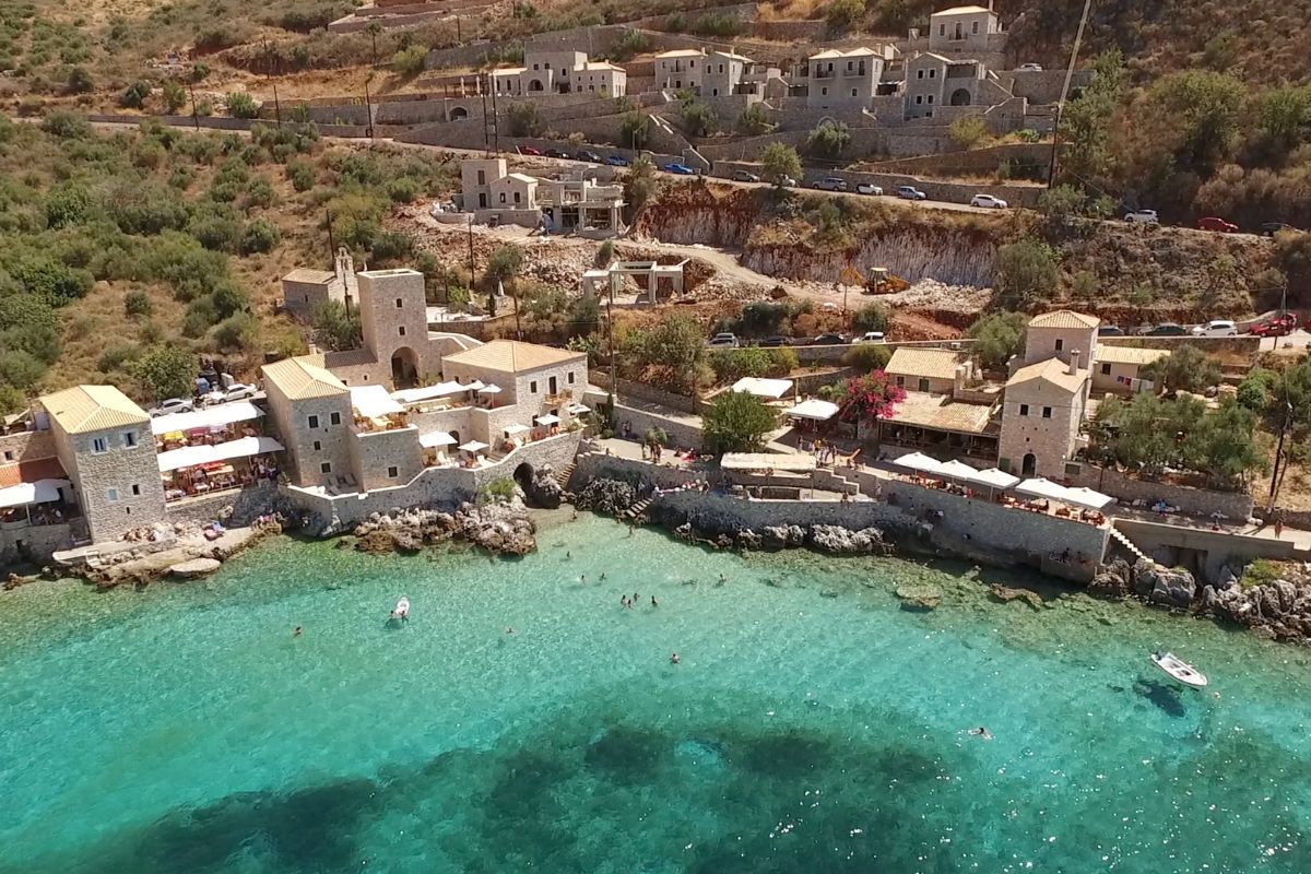 The image shows a coastal village in Mani, Peloponnese, with stone-built houses set against a hillside. The crystal-clear turquoise waters of the sea border the village, where people can be seen swimming and enjoying the peaceful surroundings. The architecture blends harmoniously with the natural landscape, creating a serene Mediterranean atmosphere.