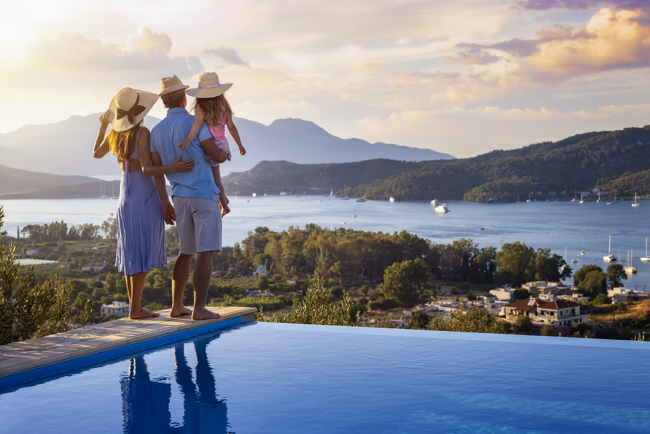 A family on summer holidays stands by the swimming pool and enjoys the beautiful sunset behind the mountains and the sea