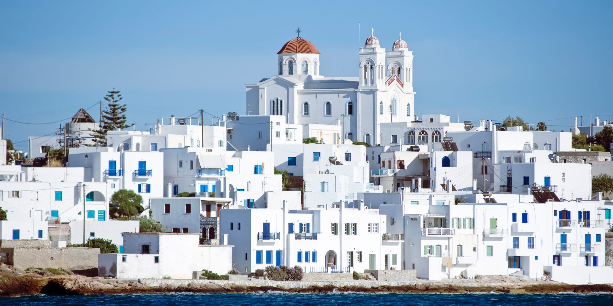 A scenic view of Paros Island featuring traditional whitewashed buildings with blue accents, and a prominent church with a red dome in the background, all set against a clear blue sky.