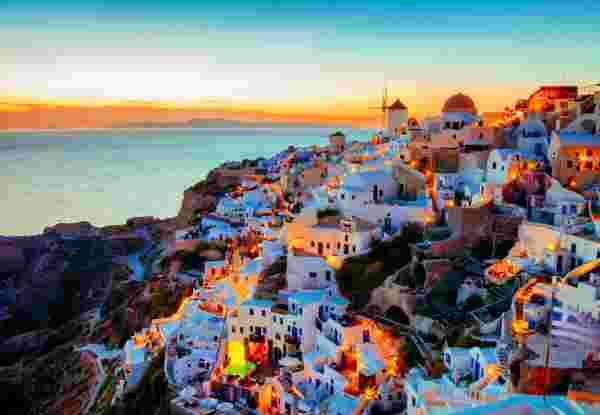 A stunning view of Santorini Island at sunset, showcasing whitewashed buildings cascading down the hillside, illuminated by warm lights, with the Aegean Sea in the background. santorini island_one day in santorini_feature