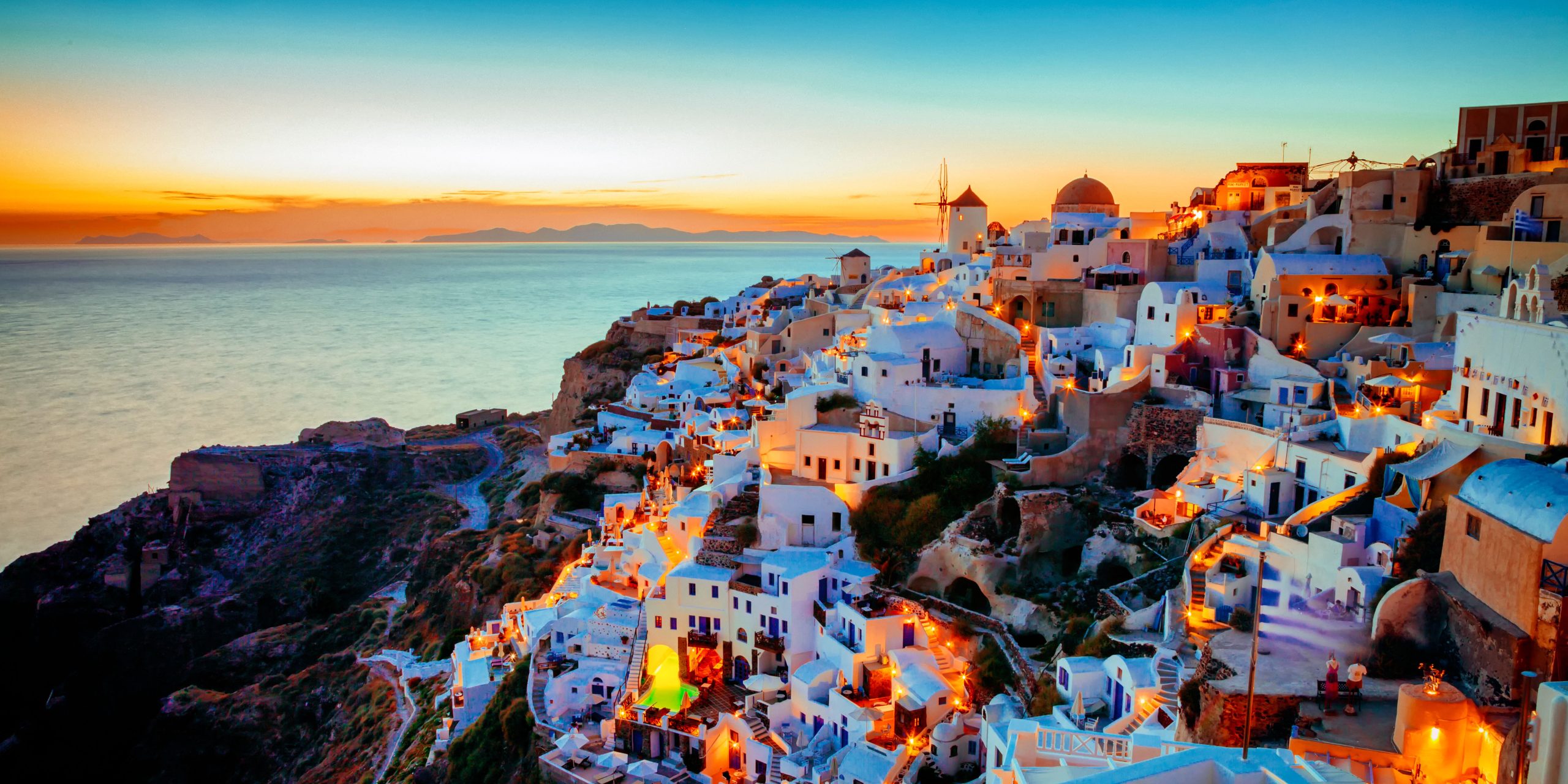 A stunning view of Santorini Island at sunset, showcasing whitewashed buildings cascading down the hillside, illuminated by warm lights, with the Aegean Sea in the background.