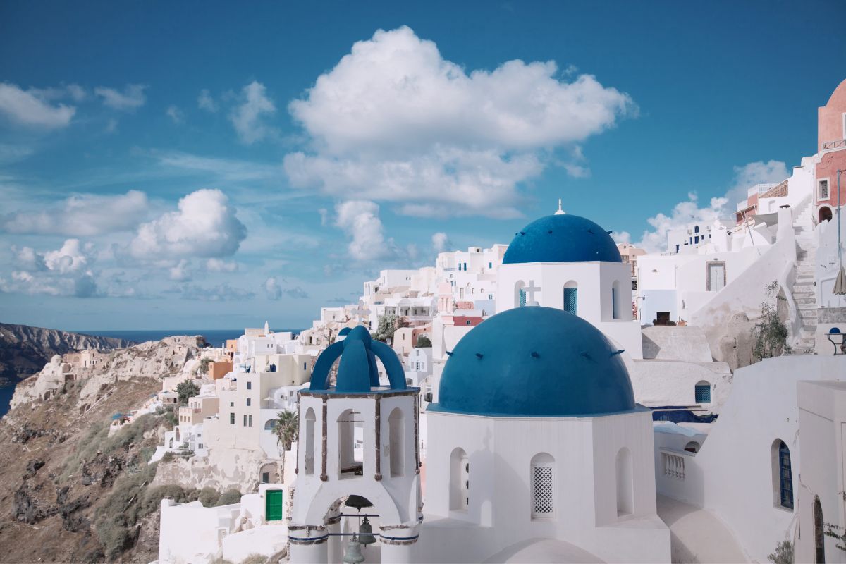 A classic view of Santorini featuring whitewashed buildings and blue-domed churches overlooking the sea. The bright blue sky and fluffy clouds add to the picturesque scene, with the island's cliffs and architecture stretching out in the background.