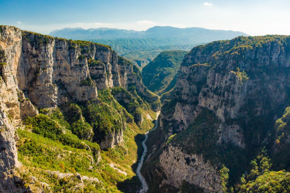 A panoramic view of Vikos Gorge, showcasing towering limestone cliffs with lush greenery along the canyon walls. A river winds through the bottom of the deep valley, with mountains visible in the distance under a clear sky.