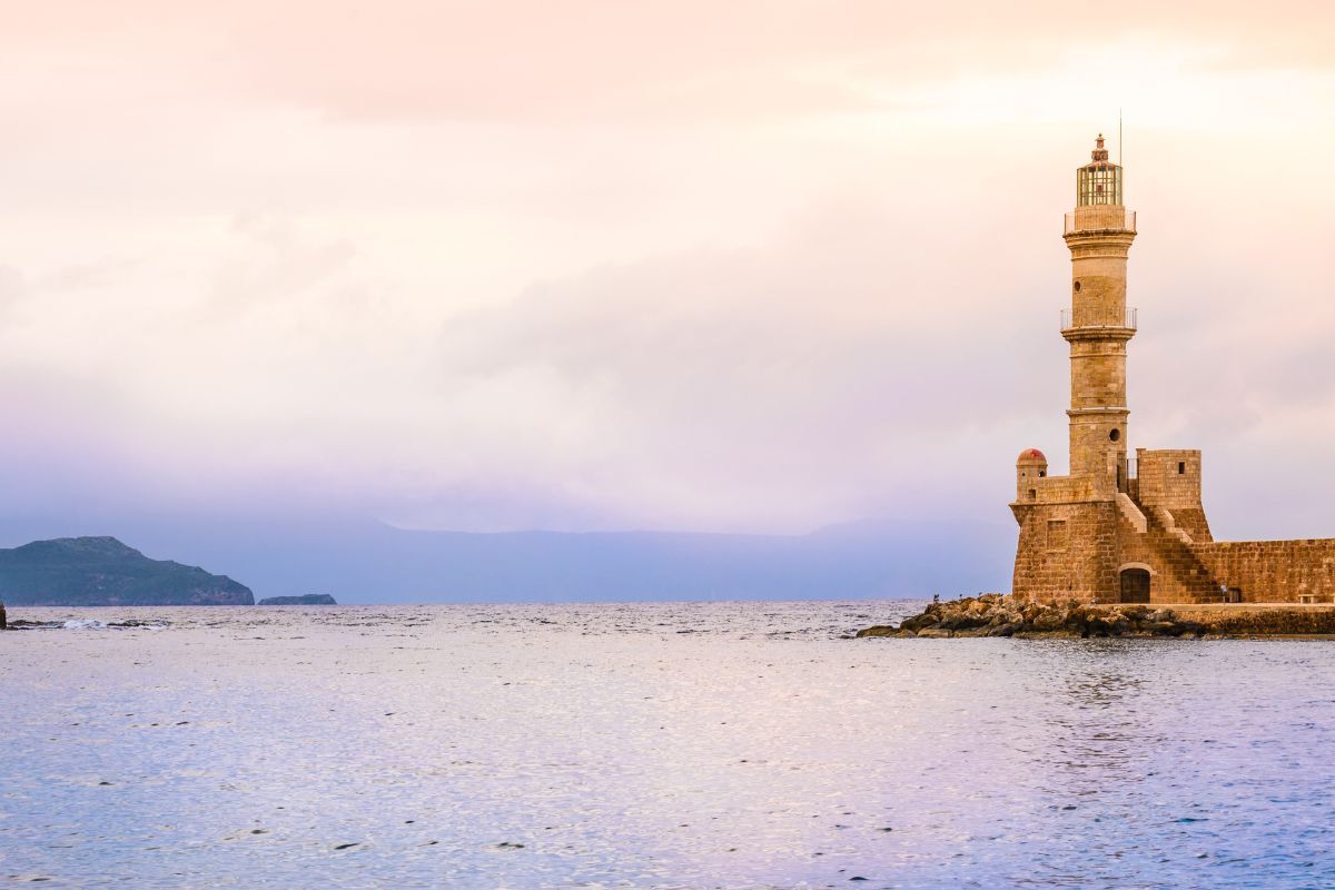 A view of the iconic lighthouse in Chania, Crete, standing at the edge of the harbor. The calm sea and soft pastel sky create a tranquil atmosphere, with distant hills visible in the background.