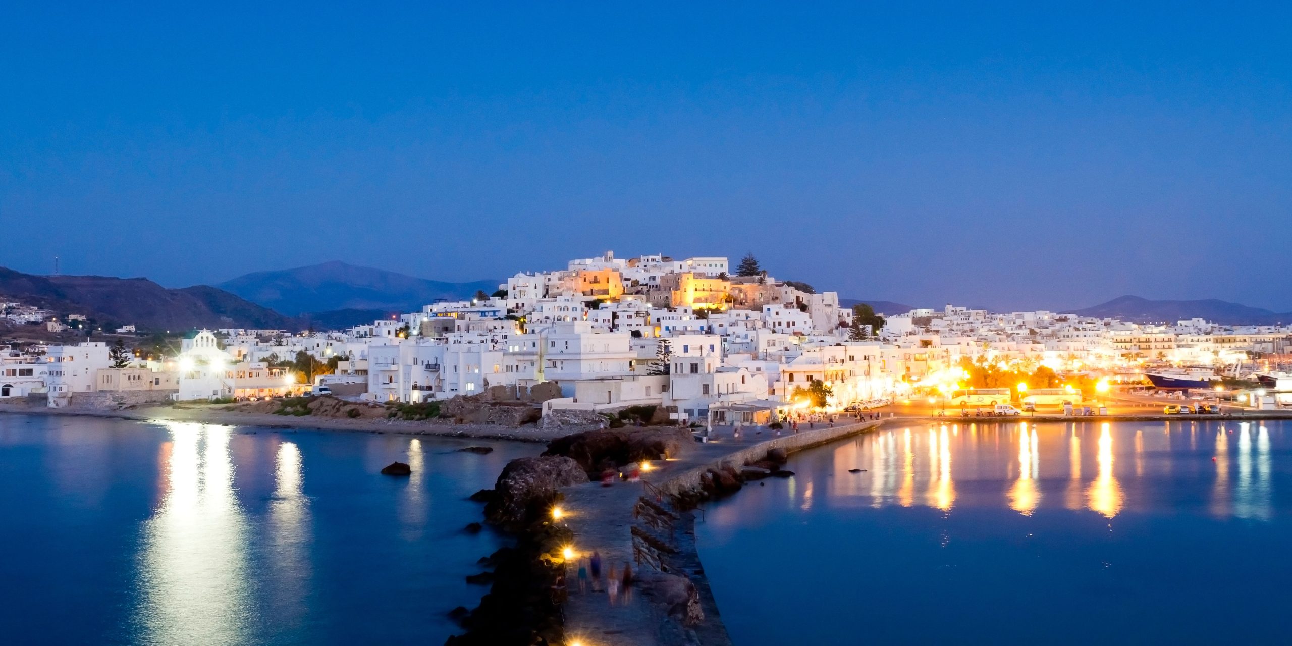 A nighttime view of Naxos, with the town's whitewashed buildings illuminated by warm lights reflecting on the calm waters. The scene captures the peaceful harbor and surrounding hills under a deep blue sky.