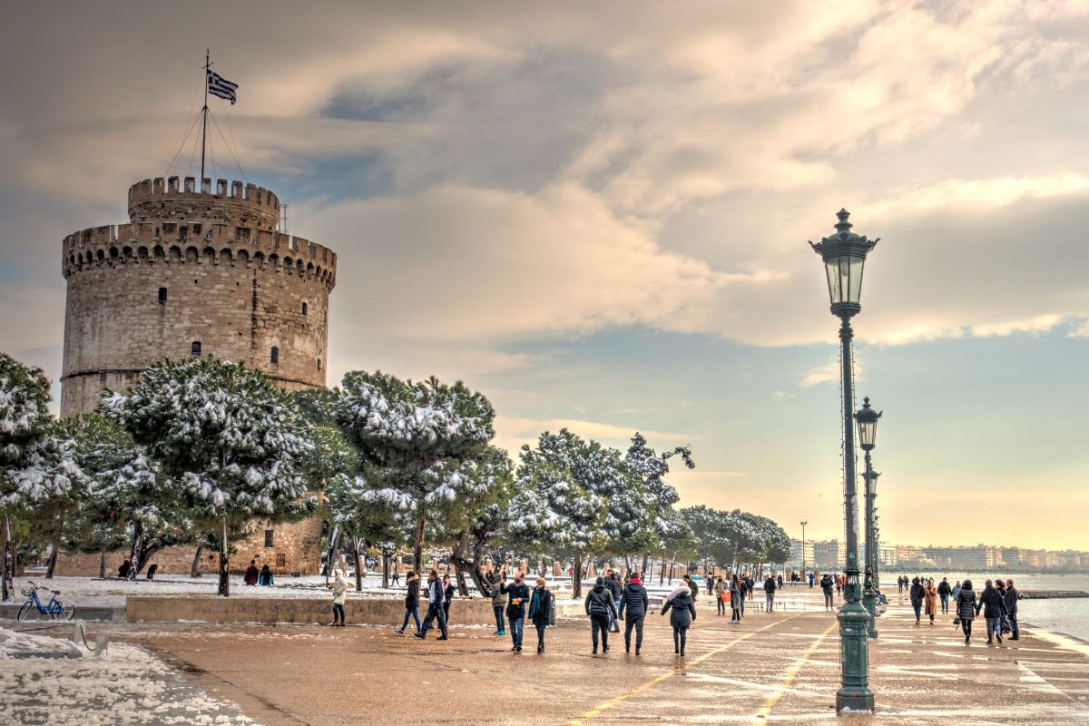 A snowy scene in Thessaloniki, featuring the iconic White Tower surrounded by snow-covered trees. People stroll along the waterfront promenade, with street lamps lining the path, while the Greek flag flies atop the tower.