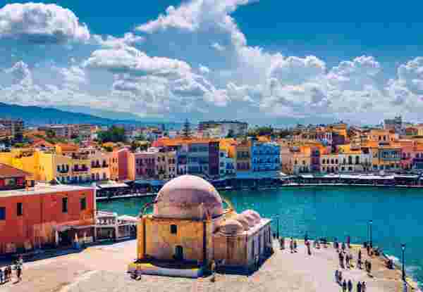 A view of Chania's Old Venetian Harbor in Crete, featuring colorful waterfront buildings, a mosque with a dome, and the sea under a partly cloudy sky. Chania Crete