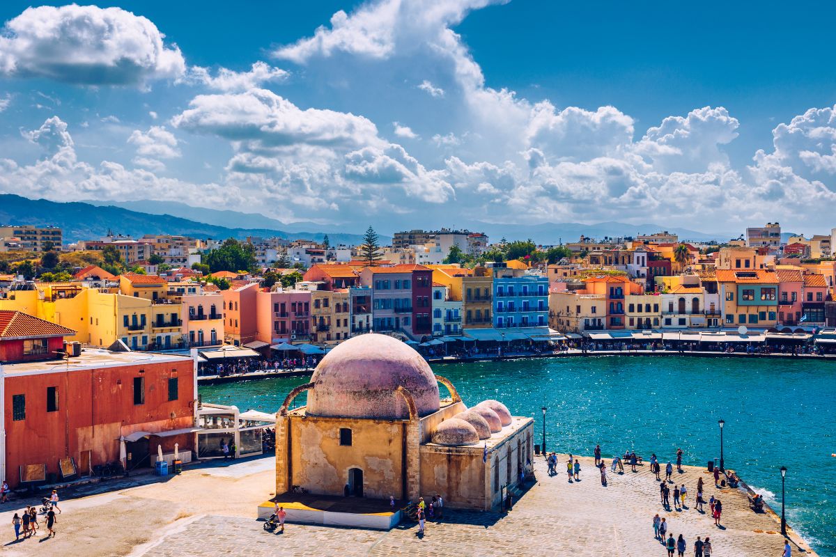 A view of Chania's Old Venetian Harbor in Crete, featuring colorful waterfront buildings, a mosque with a dome, and the sea under a partly cloudy sky.