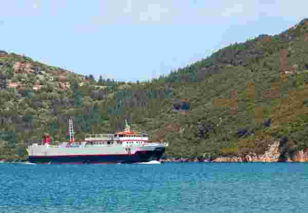 A Greek ferry sailing across calm blue waters, with a backdrop of green, forested hills. greek ferry