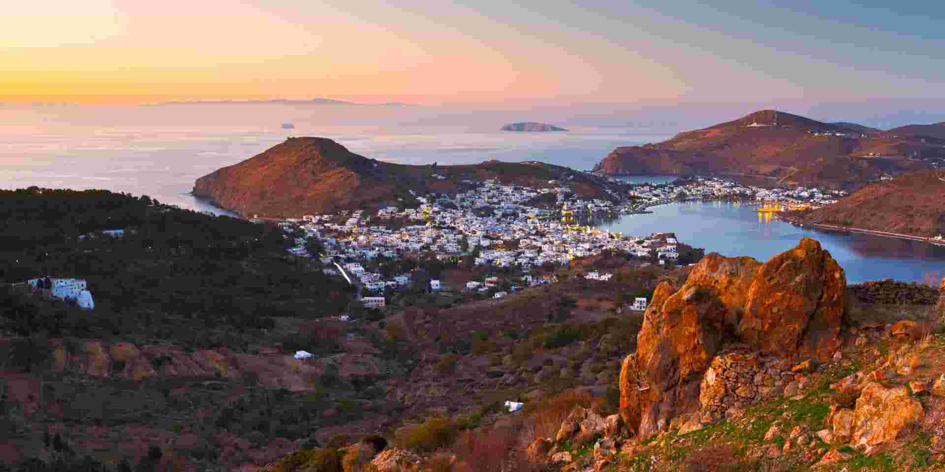 A sunset view of Patmos with a bay surrounded by hills and a town of whitewashed buildings. The calm sea reflects the soft evening light, and the rugged terrain adds depth to the landscape. Patmos_quiet greek islands_feature
