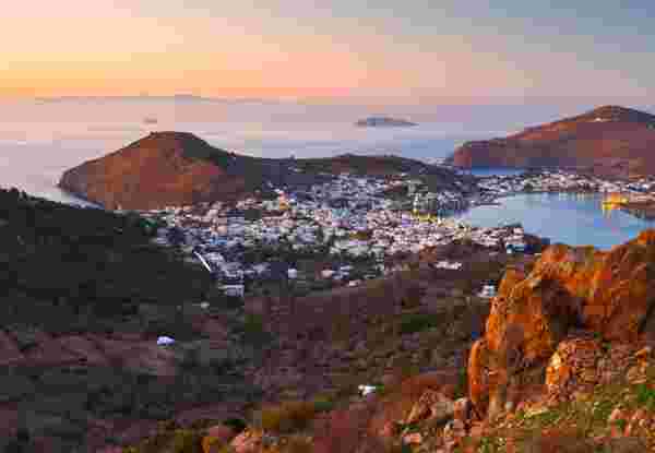 A sunset view of Patmos with a bay surrounded by hills and a town of whitewashed buildings. The calm sea reflects the soft evening light, and the rugged terrain adds depth to the landscape. Patmos_quiet greek islands_feature